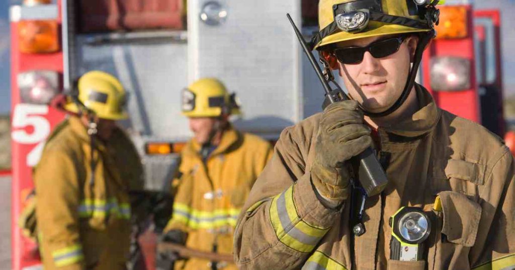 A firefighter dressed in his gear talks on a two-way radio and stands in front of a fire truck and two other firefighters.