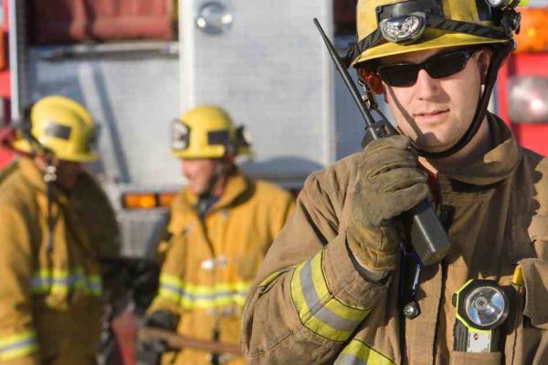 A firefighter dressed in his gear talks on a two-way radio and stands in front of a fire truck and two other firefighters.