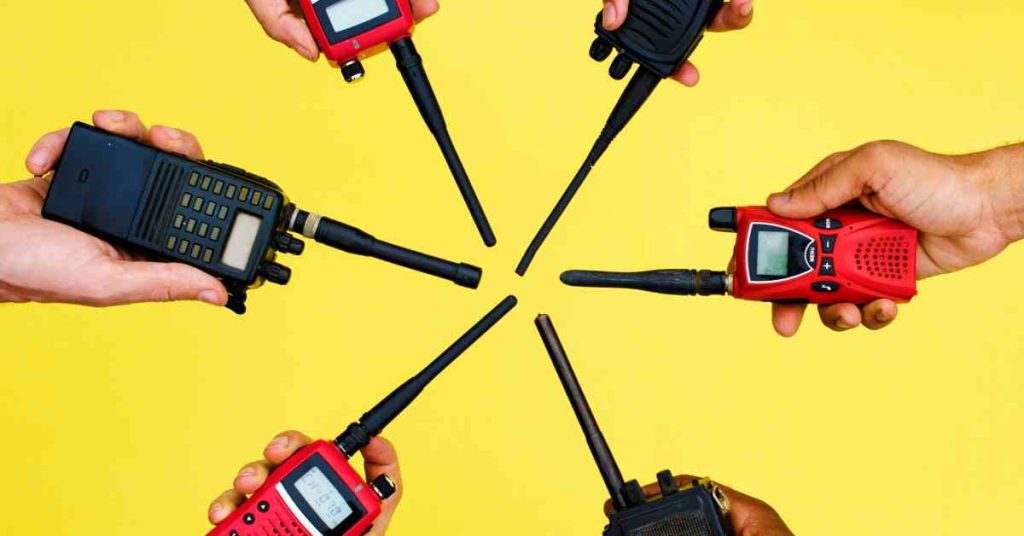 Closeup of a group of six people’s hands in a circle holding red and black two-way radios against a yellow background.