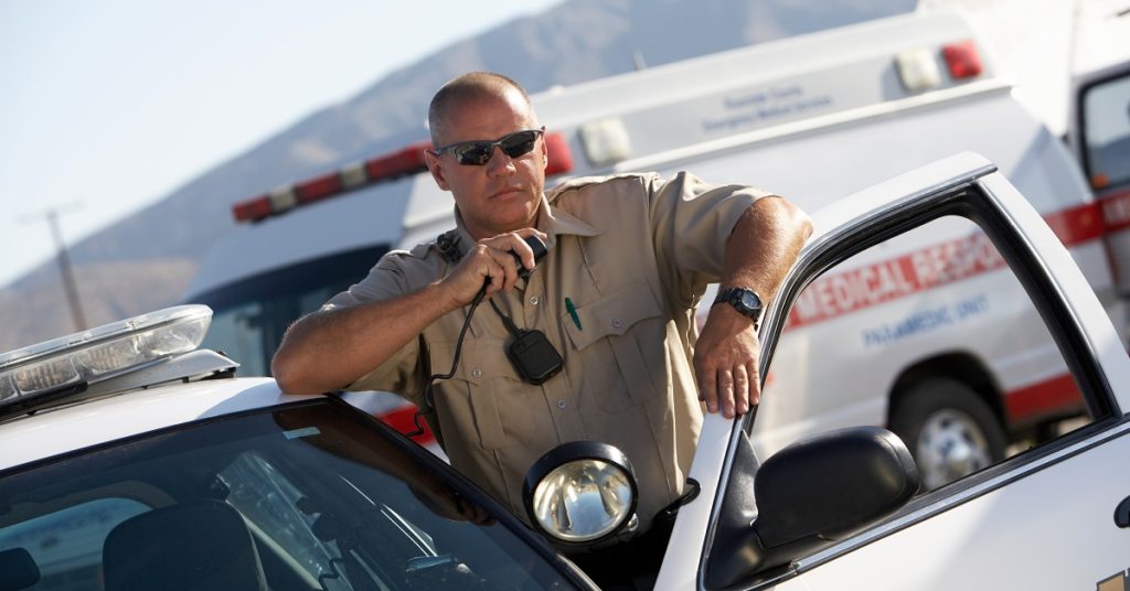 A uniformed police officer using a two-way radio as he steps out of his car. There is an ambulance in the background.