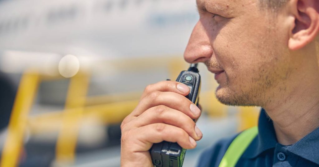 A man wearing a high-visibility vest holds a two-way radio up to his mouth in front of a blurry background.