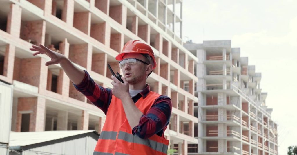  A man wearing a hard hat, goggles, and a high-visibility vest points forward while talking on a two-way radio.