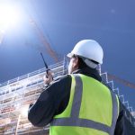 A construction worker wearing a high-visibility vest is talking on a two-way radio while looking up at a large structure.