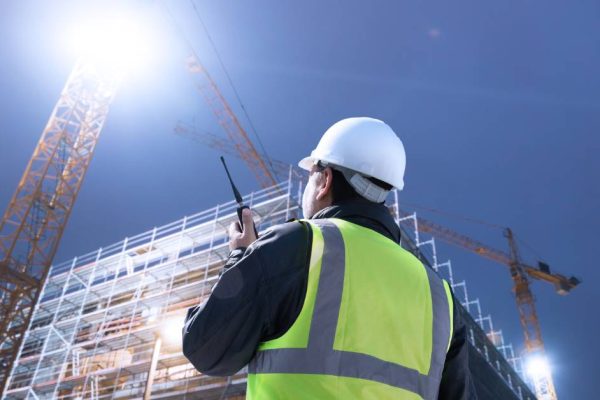 A construction worker wearing a high-visibility vest is talking on a two-way radio while looking up at a large structure.