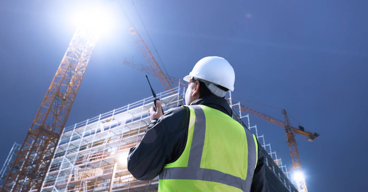 A construction worker wearing a high-visibility vest is talking on a two-way radio while looking up at a large structure.