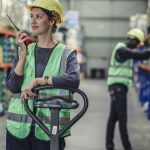 Two crew members wearing yellow hard hats and high-visibility vests in a warehouse. One is talking on a two-way radio.