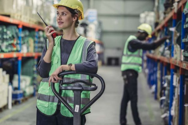 Two crew members wearing yellow hard hats and high-visibility vests in a warehouse. One is talking on a two-way radio.