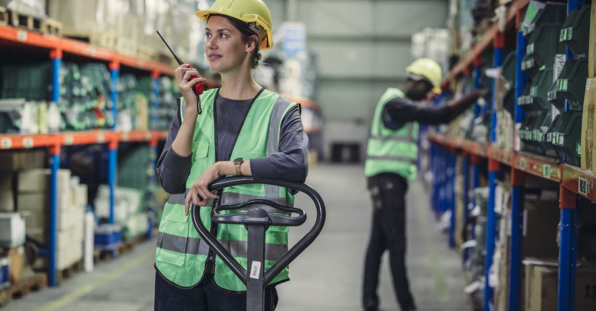 Two crew members wearing yellow hard hats and high-visibility vests in a warehouse. One is talking on a two-way radio.