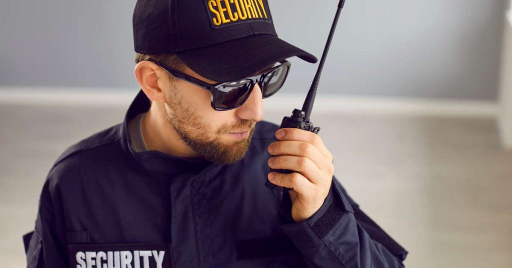 A man in a building wearing a jacket and hat that say security while holding a two-way radio and talking into it.