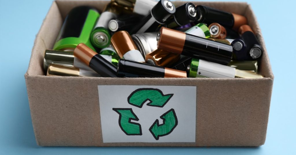 A box filled with various types of battery cells with a recycling symbol drawn on the outside of it over a blue background.