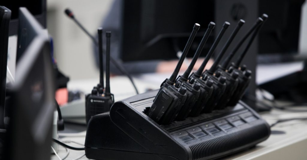 A group of black two-way radios in a charger bank on a table with another radio sitting behind them ready to charge.