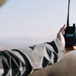 A man wearing a jacket while hiking holding a two-way radio out at arm’s length trying to pick up a signal.