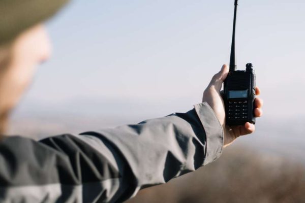 A man wearing a jacket while hiking holding a two-way radio out at arm’s length trying to pick up a signal.