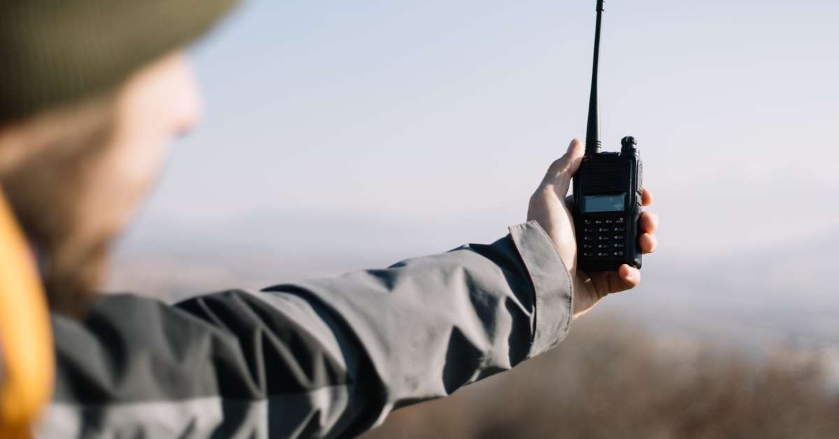 A man wearing a jacket while hiking holding a two-way radio out at arm’s length trying to pick up a signal.