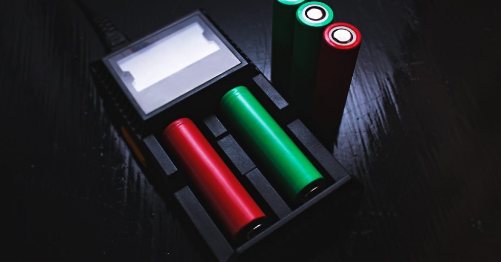 A charger with green and red battery cells. Two green batteries and one red sit upright on a table over a black background.