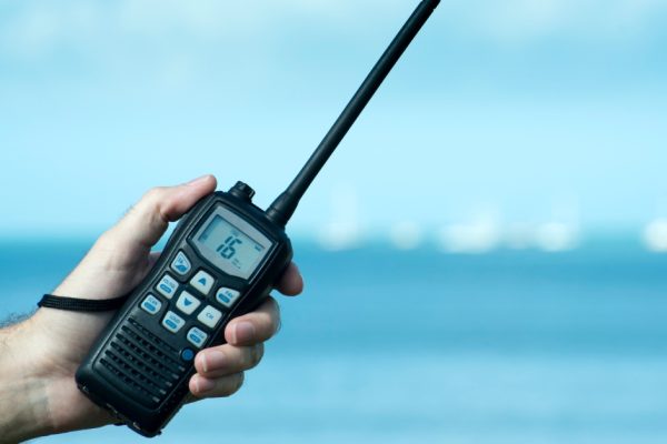 A hand holding a two-way radio in front of a body of water. The number "16" is displayed on the radio's screen.