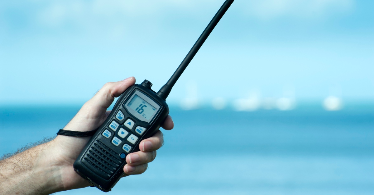 A hand holding a two-way radio in front of a body of water. The number "16" is displayed on the radio's screen.