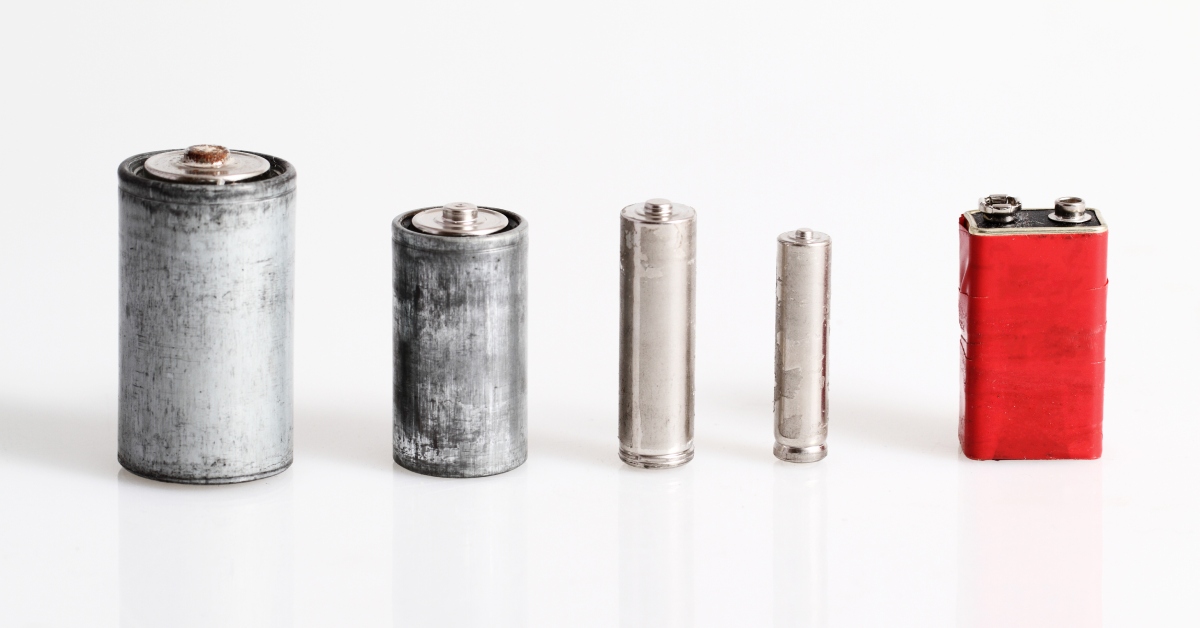 A row of large and small batteries with corrosion on the outside of the cells against a white background.