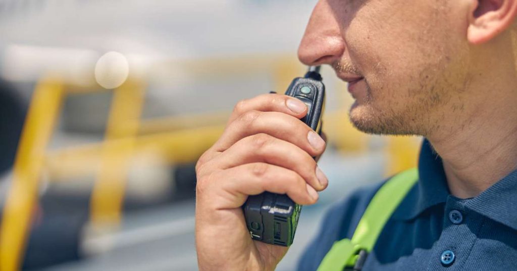 A man wearing a high-visibility vest standing outside at a worksite talking on a two-way radio he’s holding in his hand.