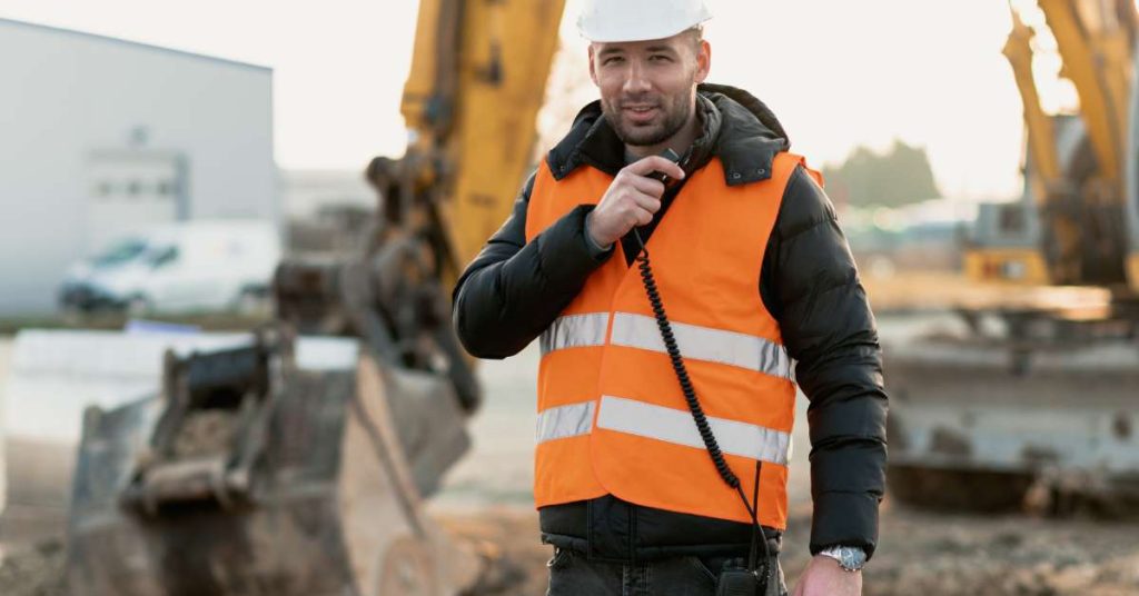 A man wearing a high-visibility vest and white hardhat talking on a two-way radio that’s clipped to his belt.