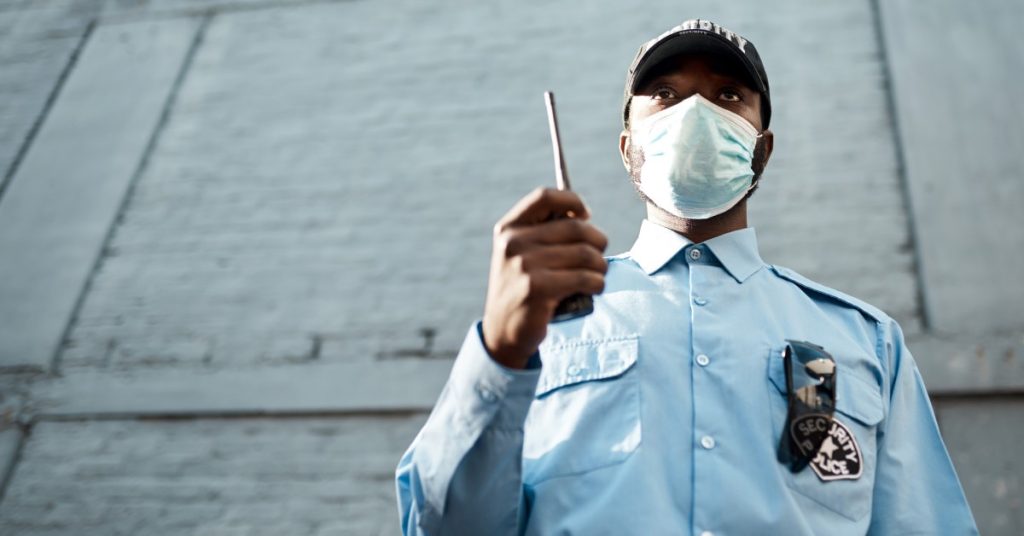 A security guard wearing a facemask standing in front of a building with a two-way radio in his hand.