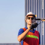 A worker in a shipping yard with a white hard hat, sunglasses, and high-visibility vest talking on a two-way radio.