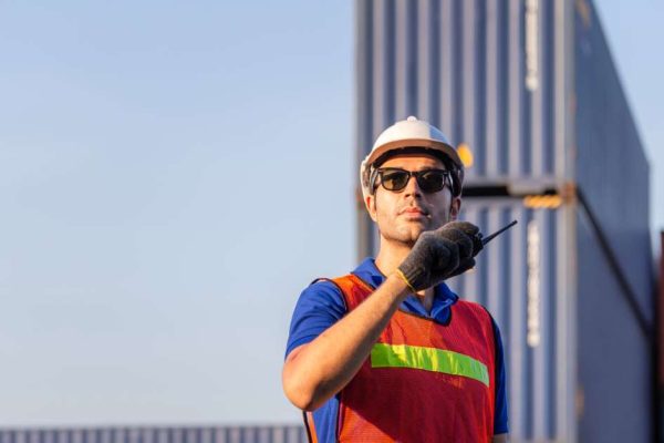 A worker in a shipping yard with a white hard hat, sunglasses, and high-visibility vest talking on a two-way radio.