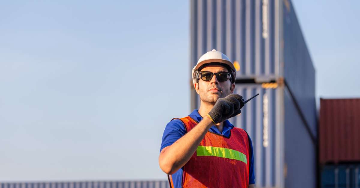 A worker in a shipping yard with a white hard hat, sunglasses, and high-visibility vest talking on a two-way radio.