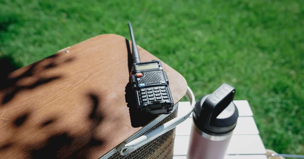 A two-way radio sitting outside on top of a table in the grass next to a thermos with the sun shining down on it.