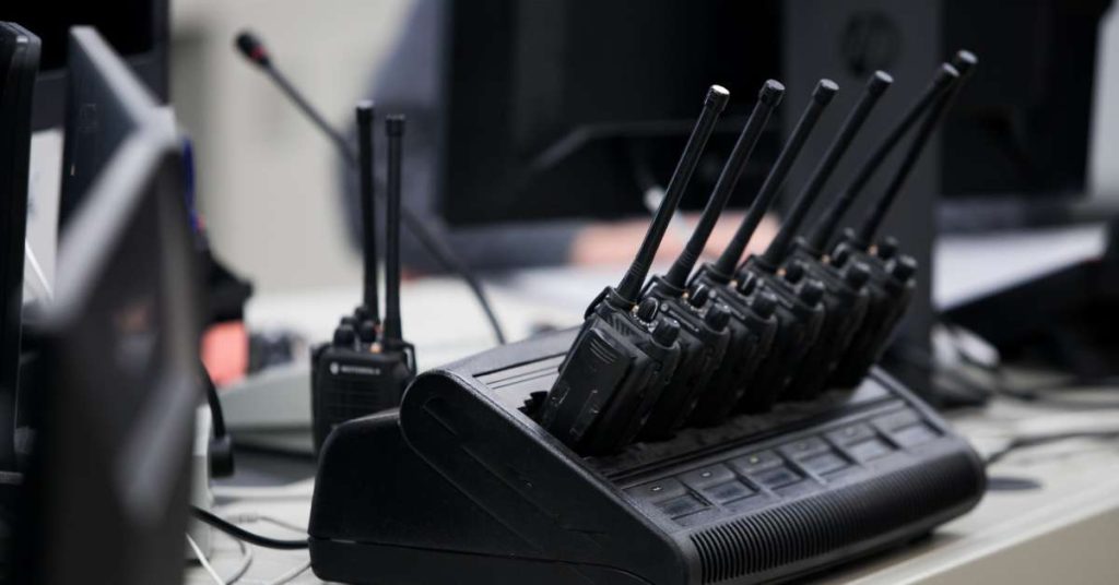 An array of two-way radios connected to a charger sitting on a desk with another radio sitting behind them.