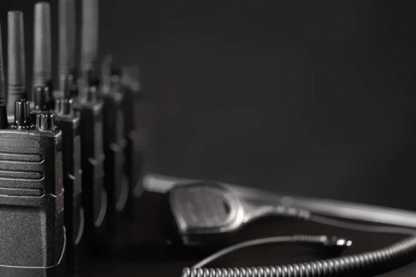 A row of five two-way radios sitting on a black table next to a wired walkie-talkie over a black background.