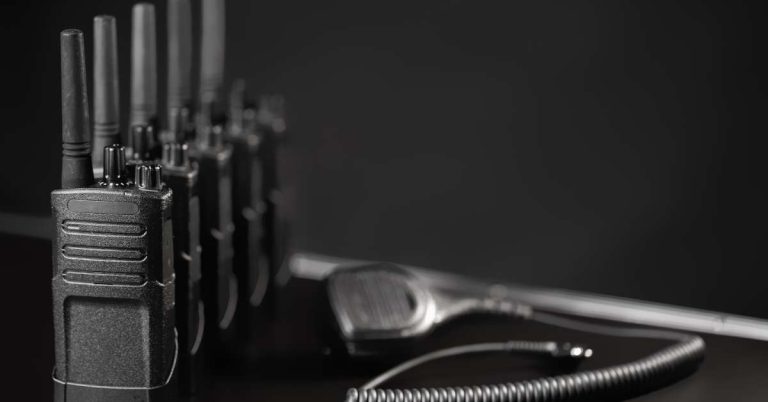 A row of five two-way radios sitting on a black table next to a wired walkie-talkie over a black background.