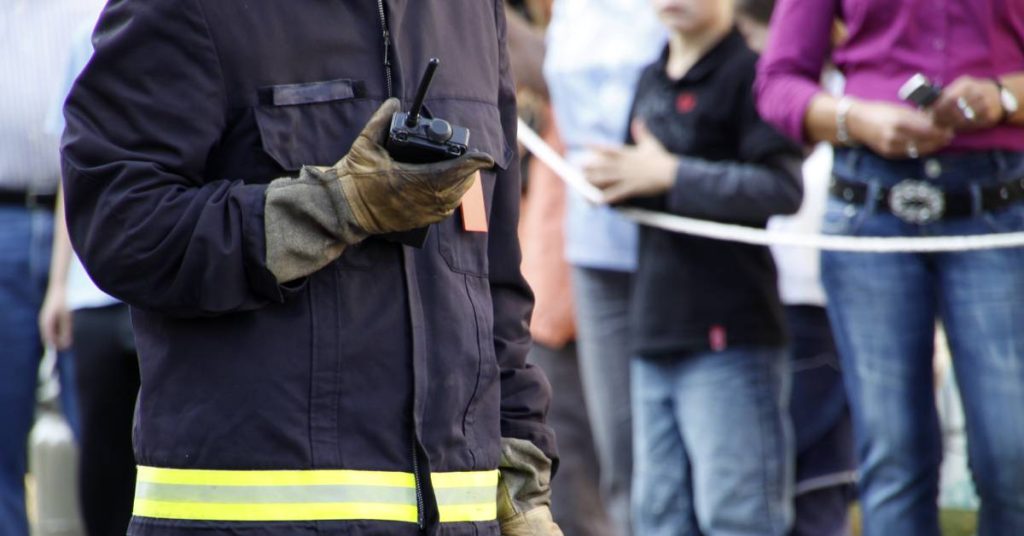 A firefighter wearing protective clothing and gloves stands in front of a crowd with a two-way radio in his hand.