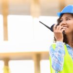 A woman wearing a blue hardhat and high-visibility vest holds a two-way radio up to her mouth with a smile.