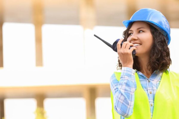 A woman wearing a blue hardhat and high-visibility vest holds a two-way radio up to her mouth with a smile.