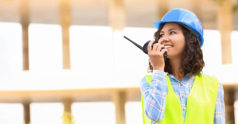 A woman wearing a blue hardhat and high-visibility vest holds a two-way radio up to her mouth with a smile.