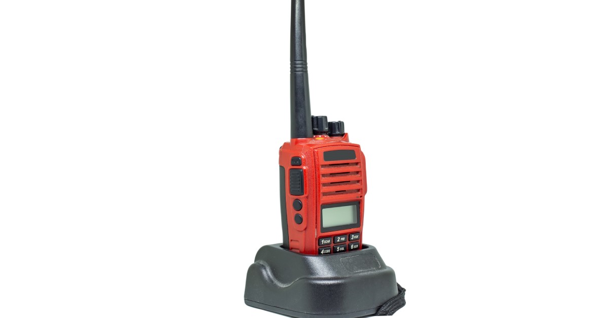 A red handheld radio with a blank screen, a black antenna, and multiple black buttons connected to a black charger.