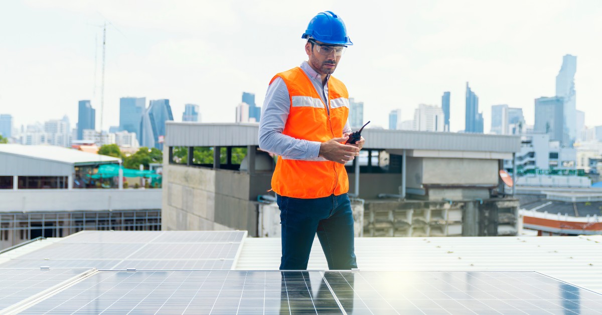 A worker in a blue hard hat and orange high-visibility vest standing in front of a solar panel while holding a two-way radio.