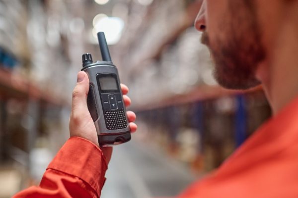 A worker in a red long-sleeve shirt holding a two-way radio close to his face. There is a blurry warehouse in the background.