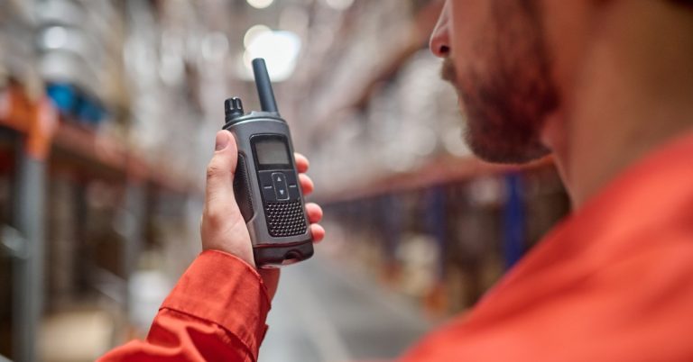 A worker in a red long-sleeve shirt holding a two-way radio close to his face. There is a blurry warehouse in the background.