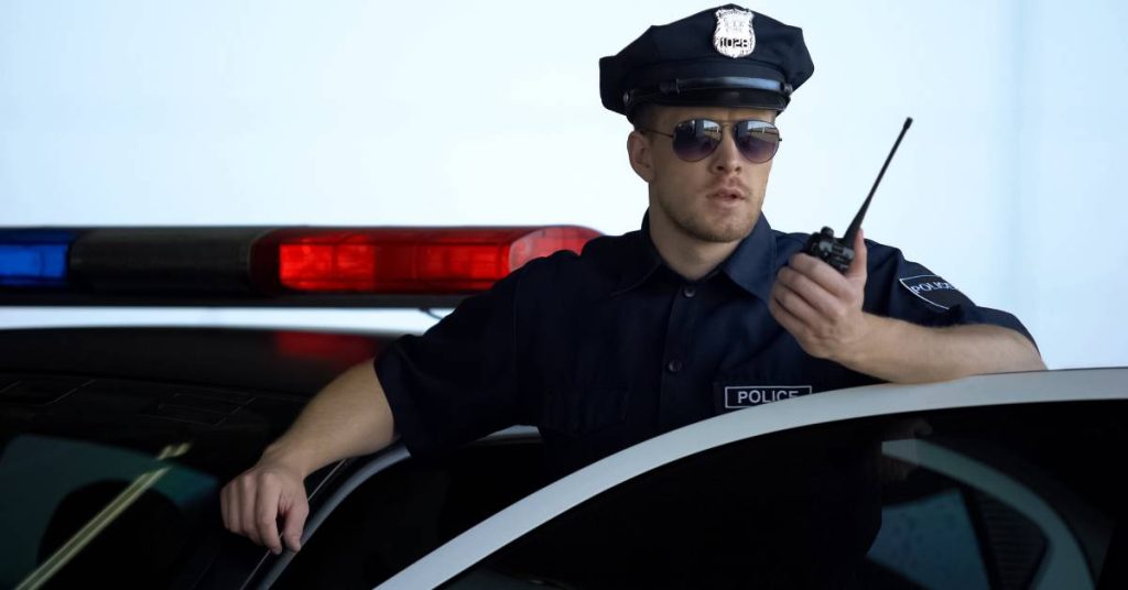 A police officer wearing sunglasses stands behind his car door and talks on a two-way radio. A bright light is in the background.