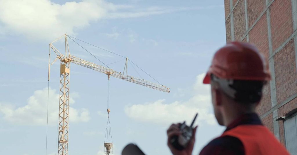 A construction worker wearing a hard hat and high visibility vest is looking at a crane while holding a two-way radio.