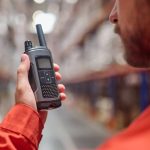 A worker wearing a long-sleeve shirt is holding a two-way radio in his hand. A warehouse floor is blurred out behind him.