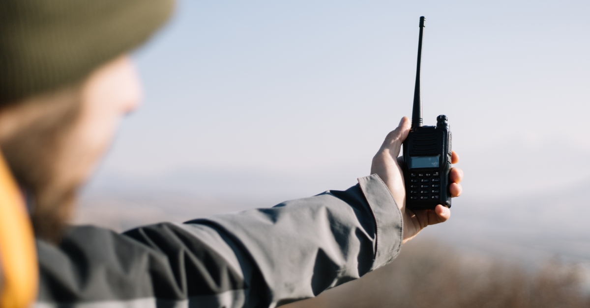 A man wearing a jacket and hat holding a two-way radio out at arm’s length in front of a blurry background.