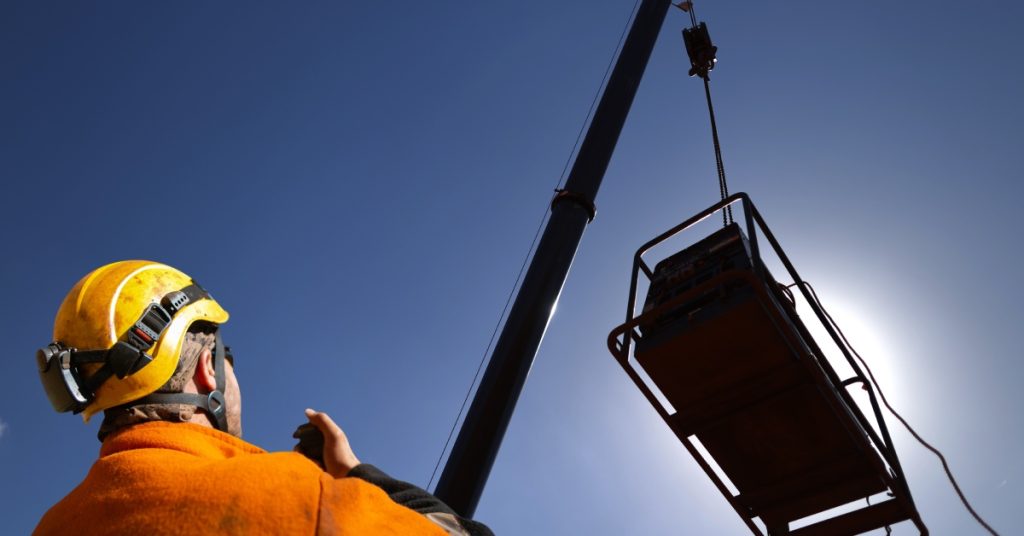 A construction worker talking into a two-way radio while looking up at a crane attached to an object with the sun behind it.