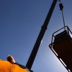 A construction worker talking into a two-way radio while looking up at a crane attached to an object with the sun behind it.