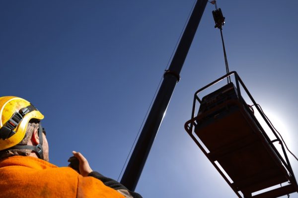 A construction worker talking into a two-way radio while looking up at a crane attached to an object with the sun behind it.