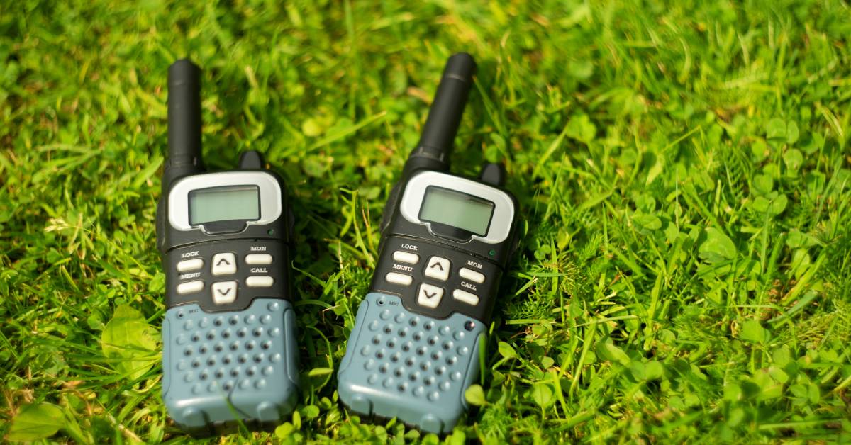 A close-up of two black two-way radios with white buttons and green speakers lying in the grass.