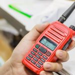 Close-up of a red two-way radio in a person's hand. In the background is a desk with stacks of paper on it.