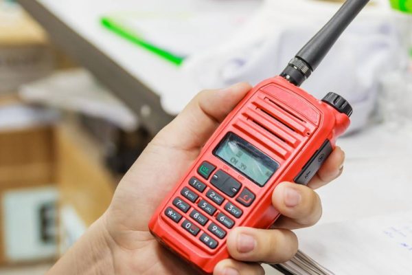 Close-up of a red two-way radio in a person's hand. In the background is a desk with stacks of paper on it.